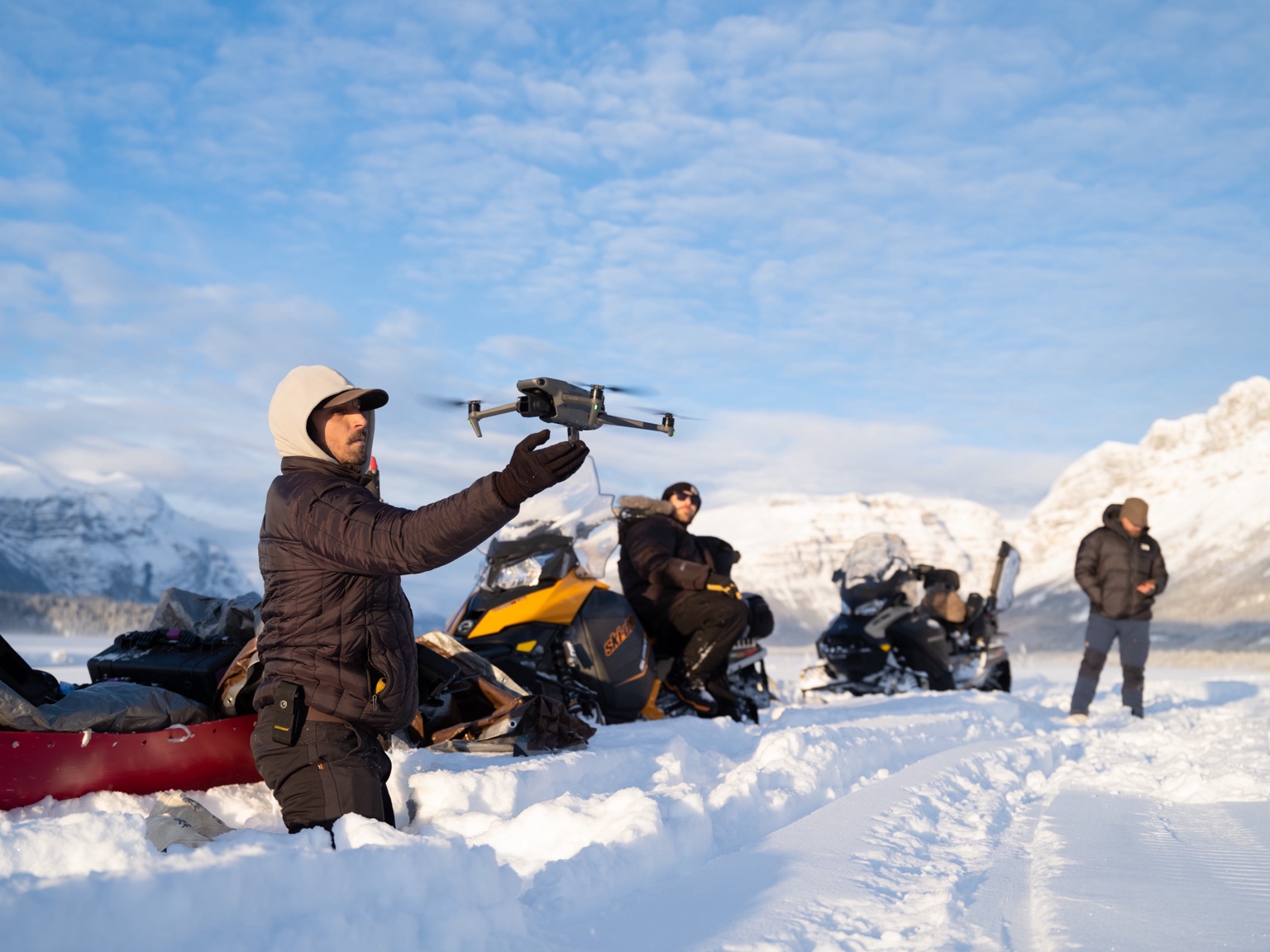 Anthony DeLeo receives a drone beside snow machines in a snowy Alaskan mountain landscape.