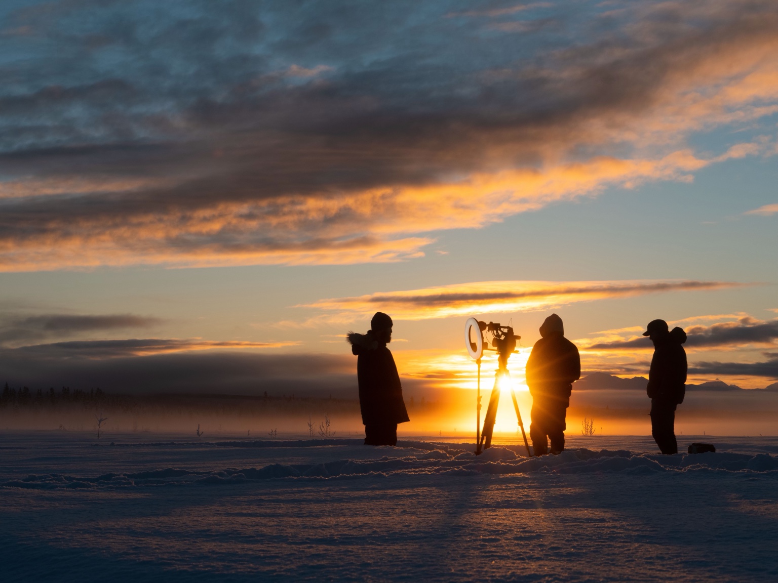 An interview crew and camera silhouetted against golden sunset light on a snowy Alaskan landscape.