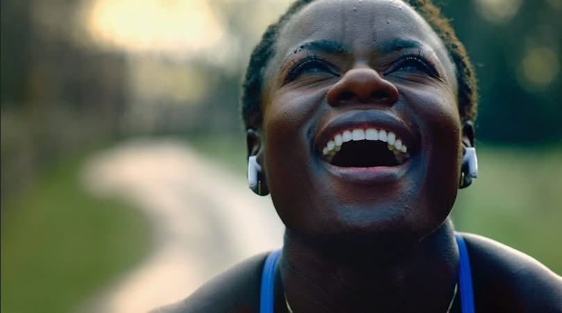 A woman wearing earbuds smiles in close-up, with a sunlit path softly out of focus behind her.