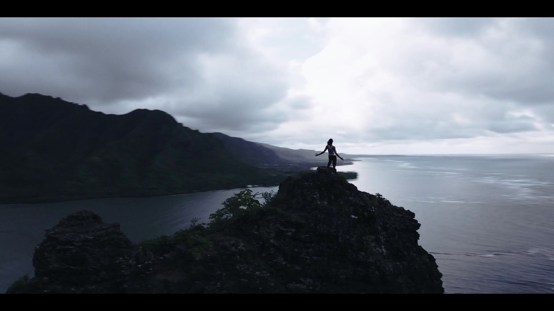 A person stands on a rocky coastal ridge above the ocean under a cloudy sky.