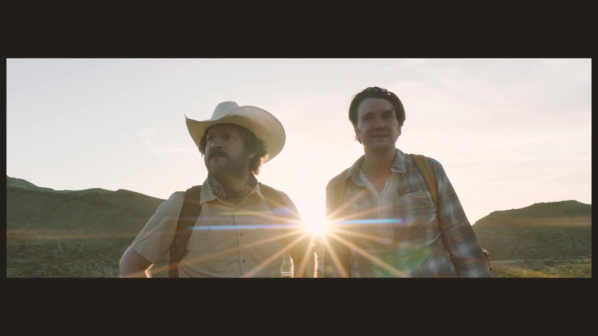 Two men stand outdoors in warm evening sunlight, with the sun flaring between them.
